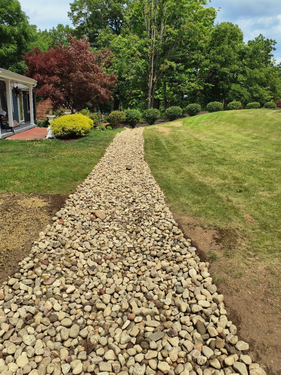 Gravel-filled dry creek bed in a residential yard. Grass surrounds it; trees and a house are in the background.