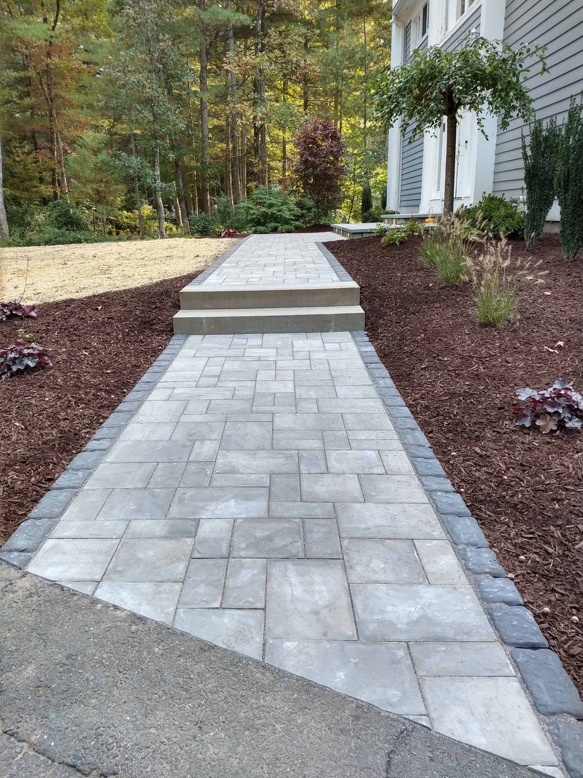 Stone walkway leading to a house, with dark edging and mulch, surrounded by trees.