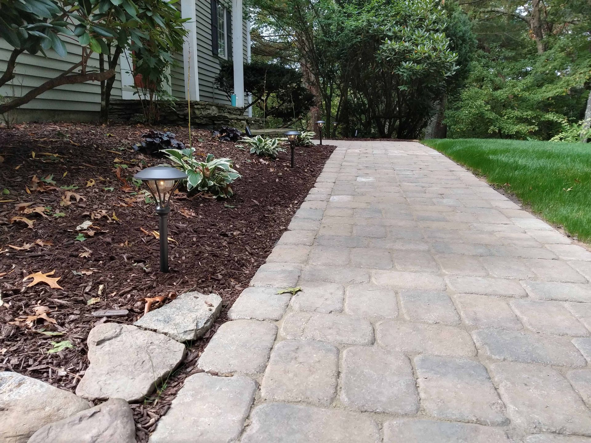 Brick walkway alongside a landscaped bed with lighting and green lawn, leading to a house.