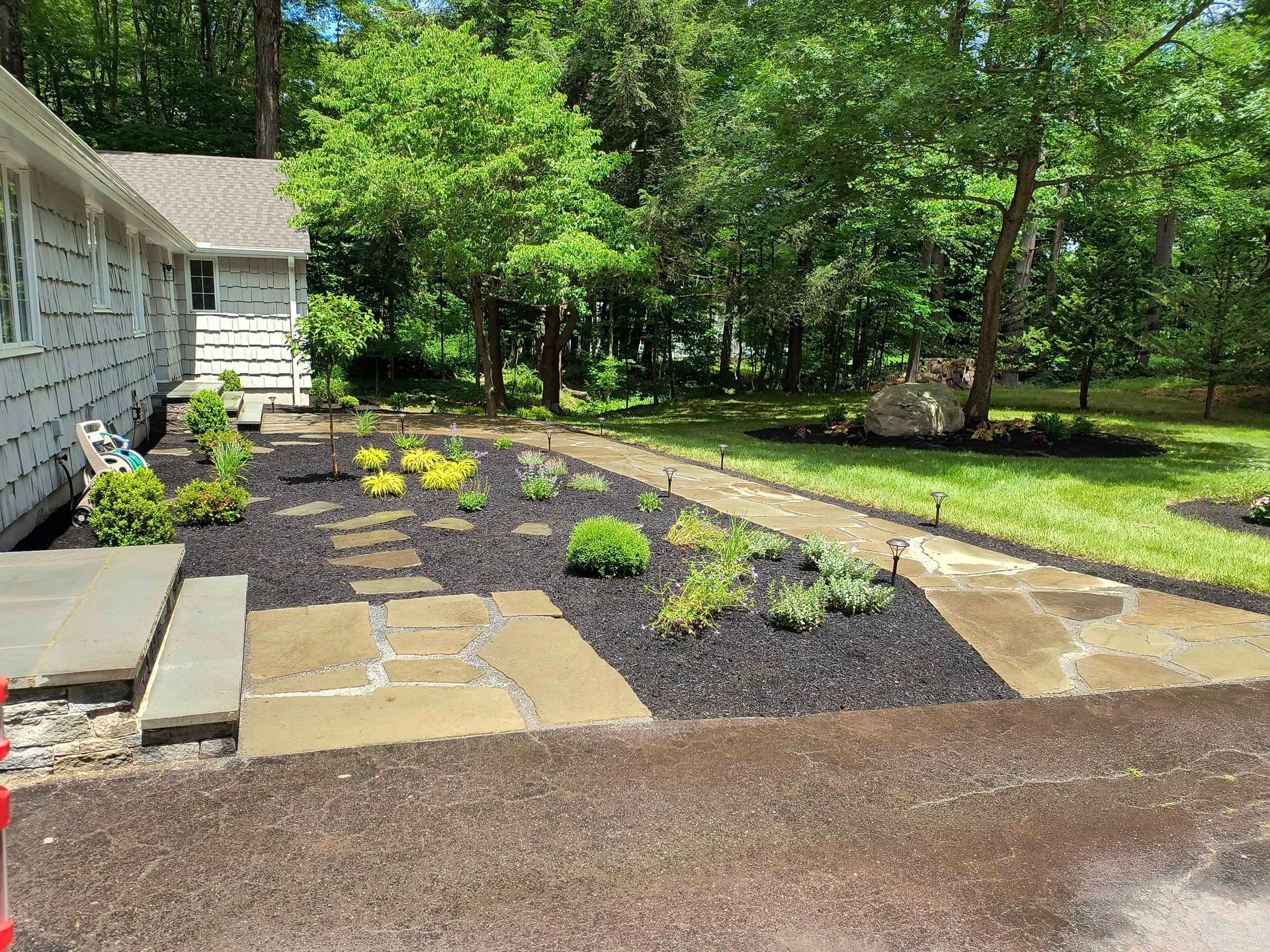 Stone path leads through a garden bed with plants and black mulch beside a house and grassy lawn.
