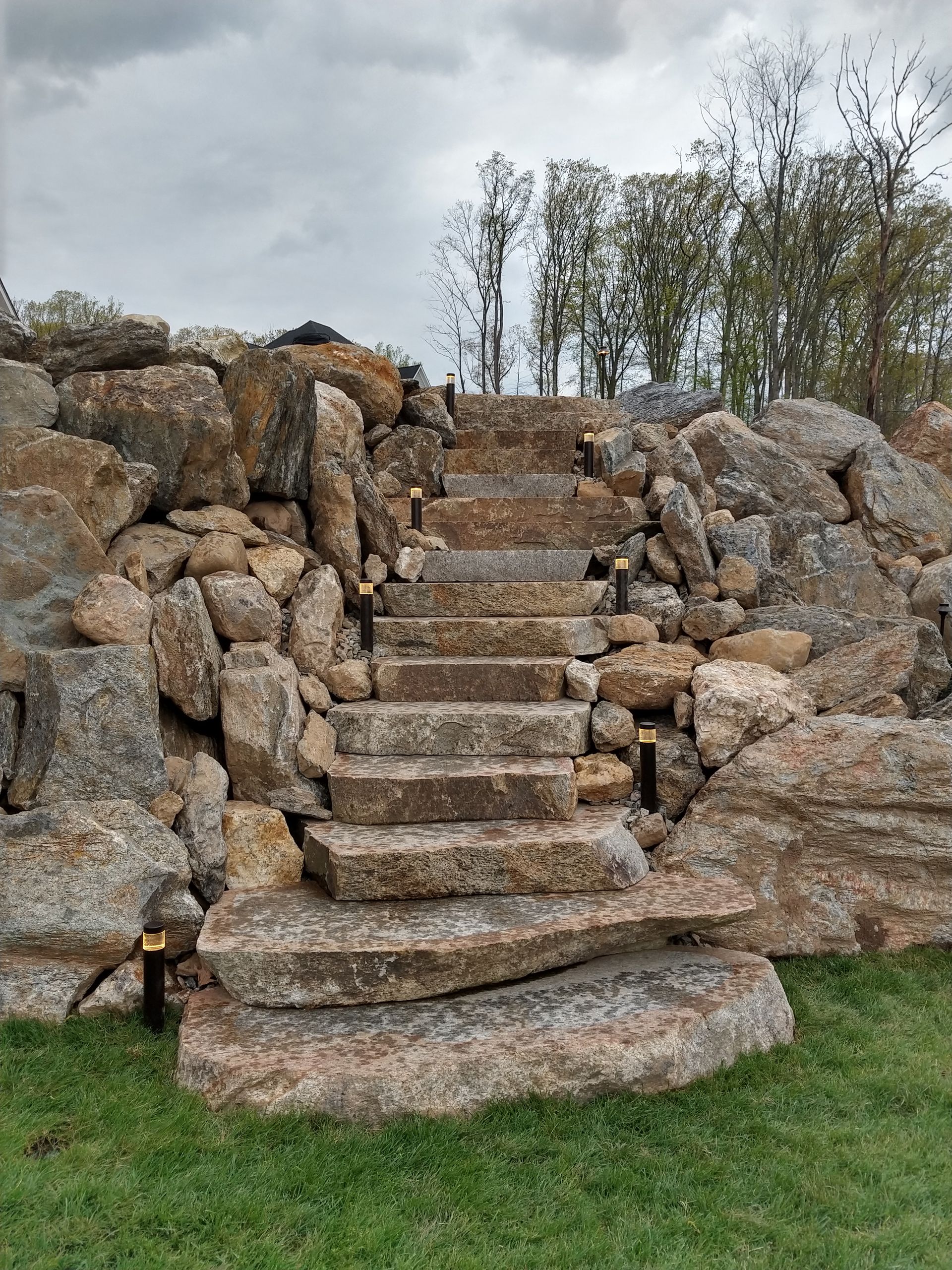 Stone steps lead up a rock wall, lit by small lights. Cloudy sky and trees in the background.