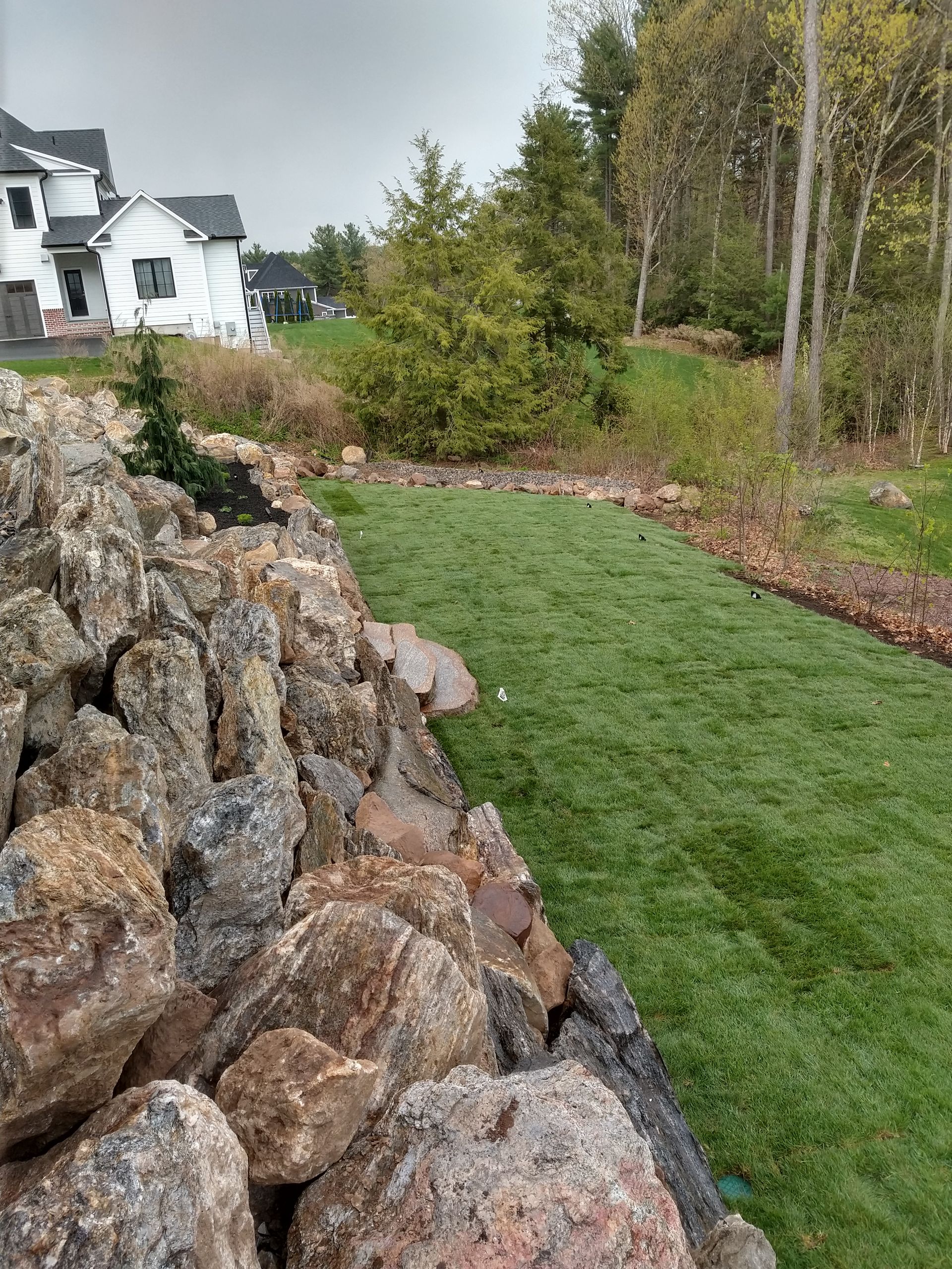 Rocky retaining wall next to a strip of green grass, with trees in the background, a house visible.