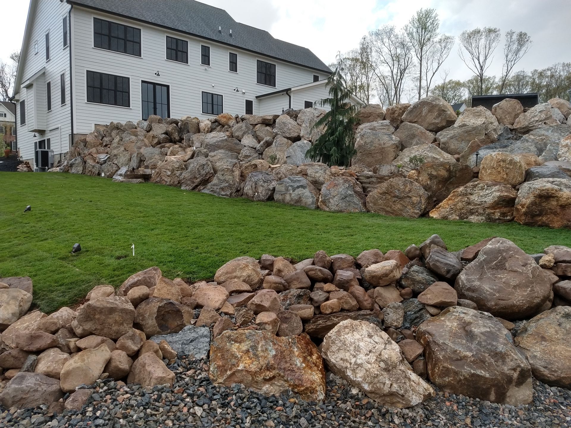Large rocks form a retaining wall in front of a white house with a green lawn.