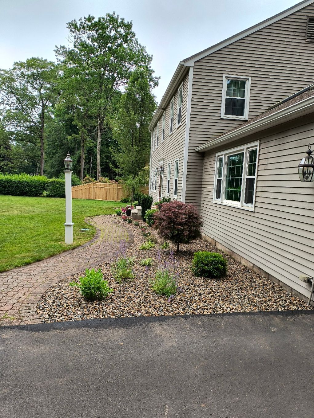 Side of a two-story beige house with landscaped garden beds, a brick path, and asphalt driveway. Overcast sky.