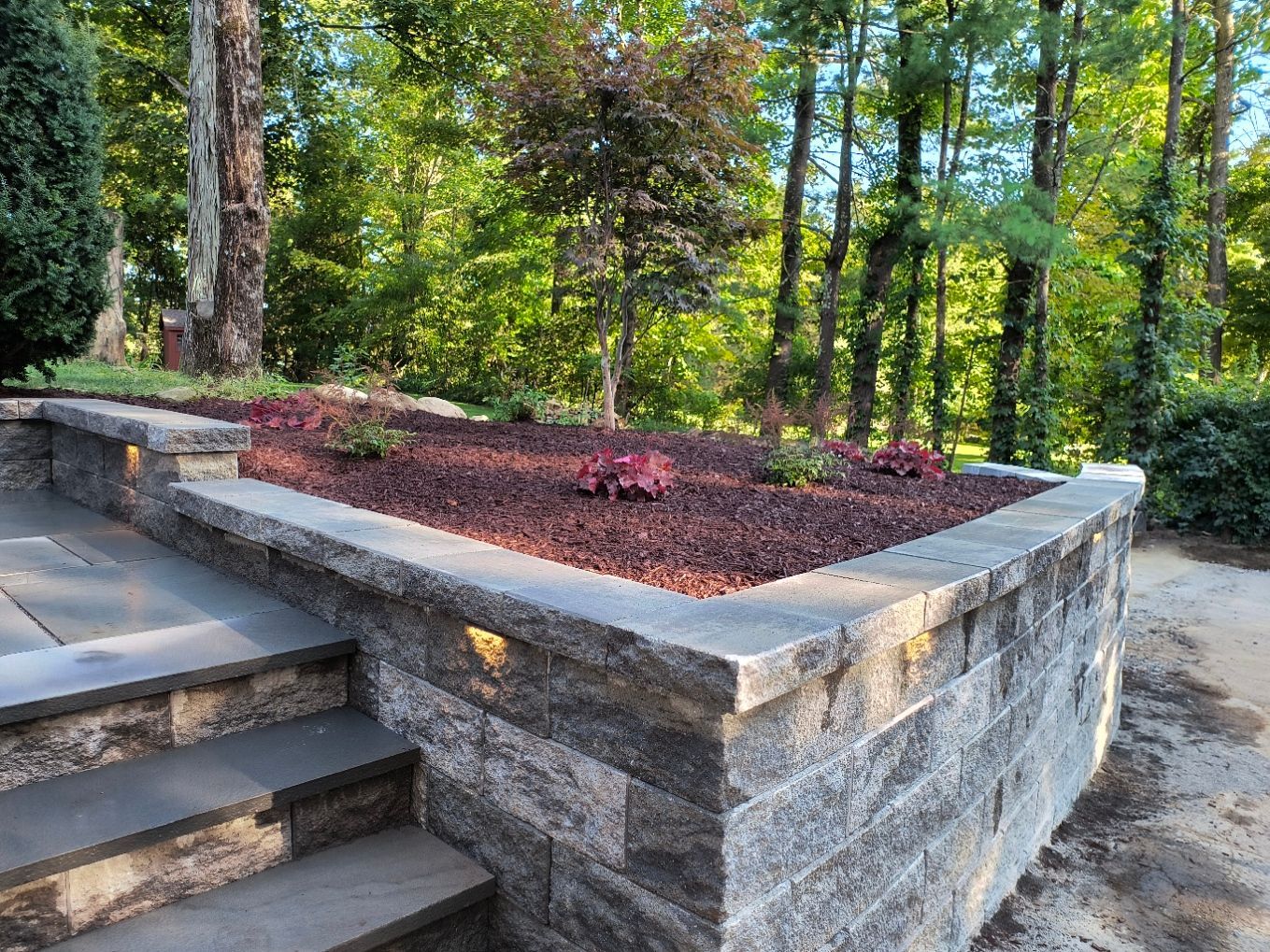 Stone retaining wall with steps leading up to a raised flowerbed filled with dark mulch and plants. Trees in background.