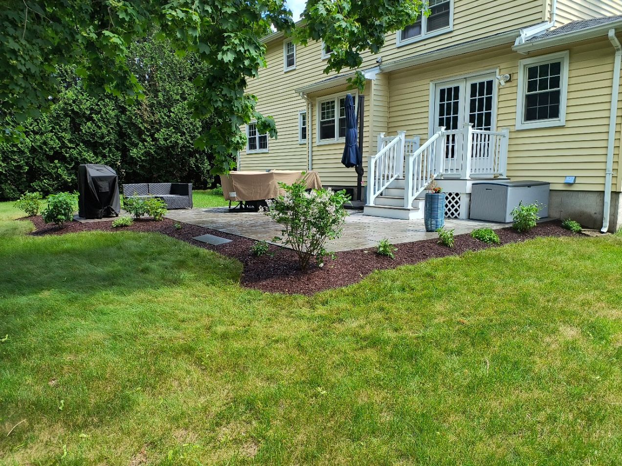 Backyard with a stone patio, landscaping, and a yellow house.