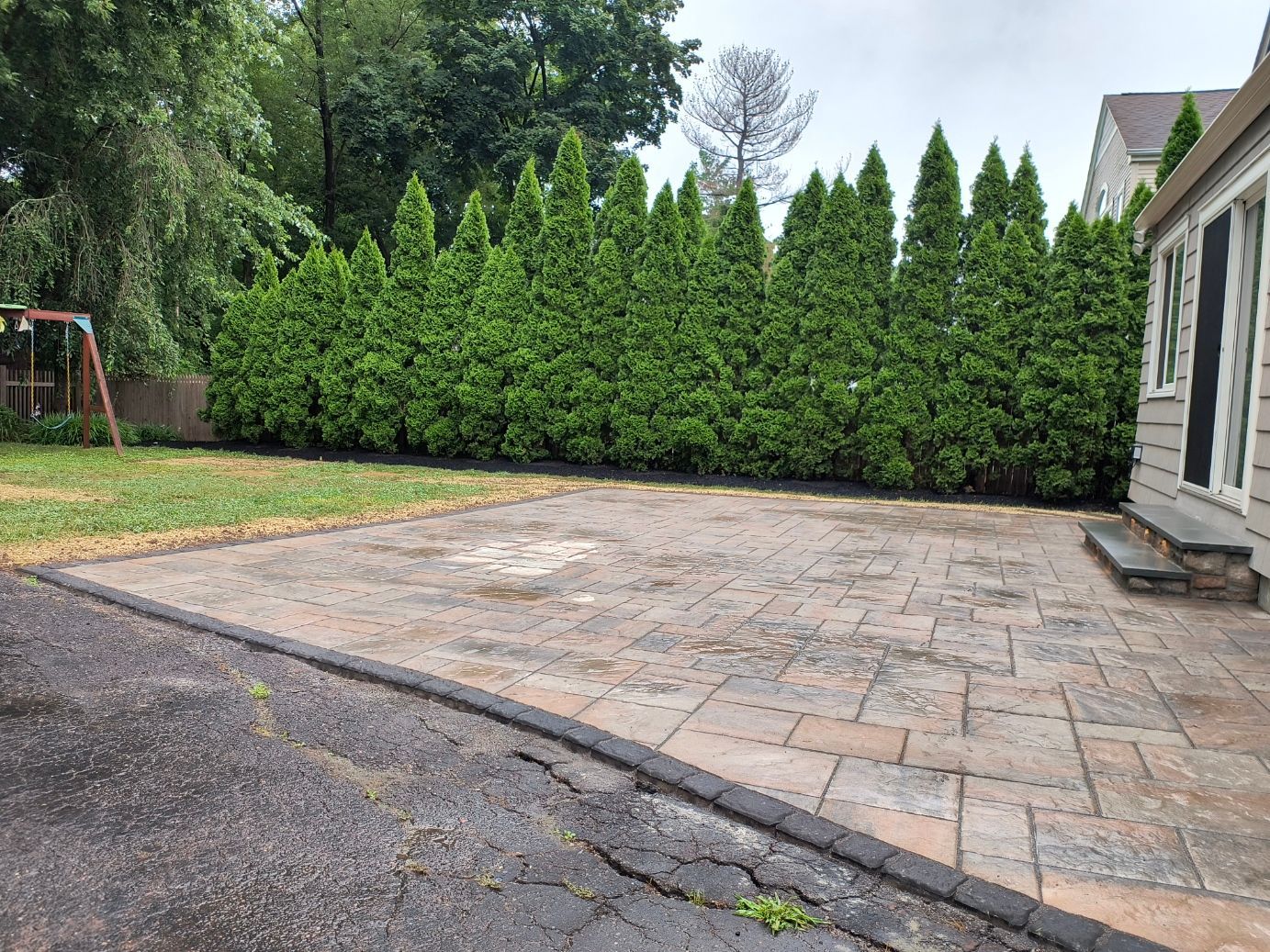 Brick patio next to a building and evergreen trees, with an asphalt driveway.
