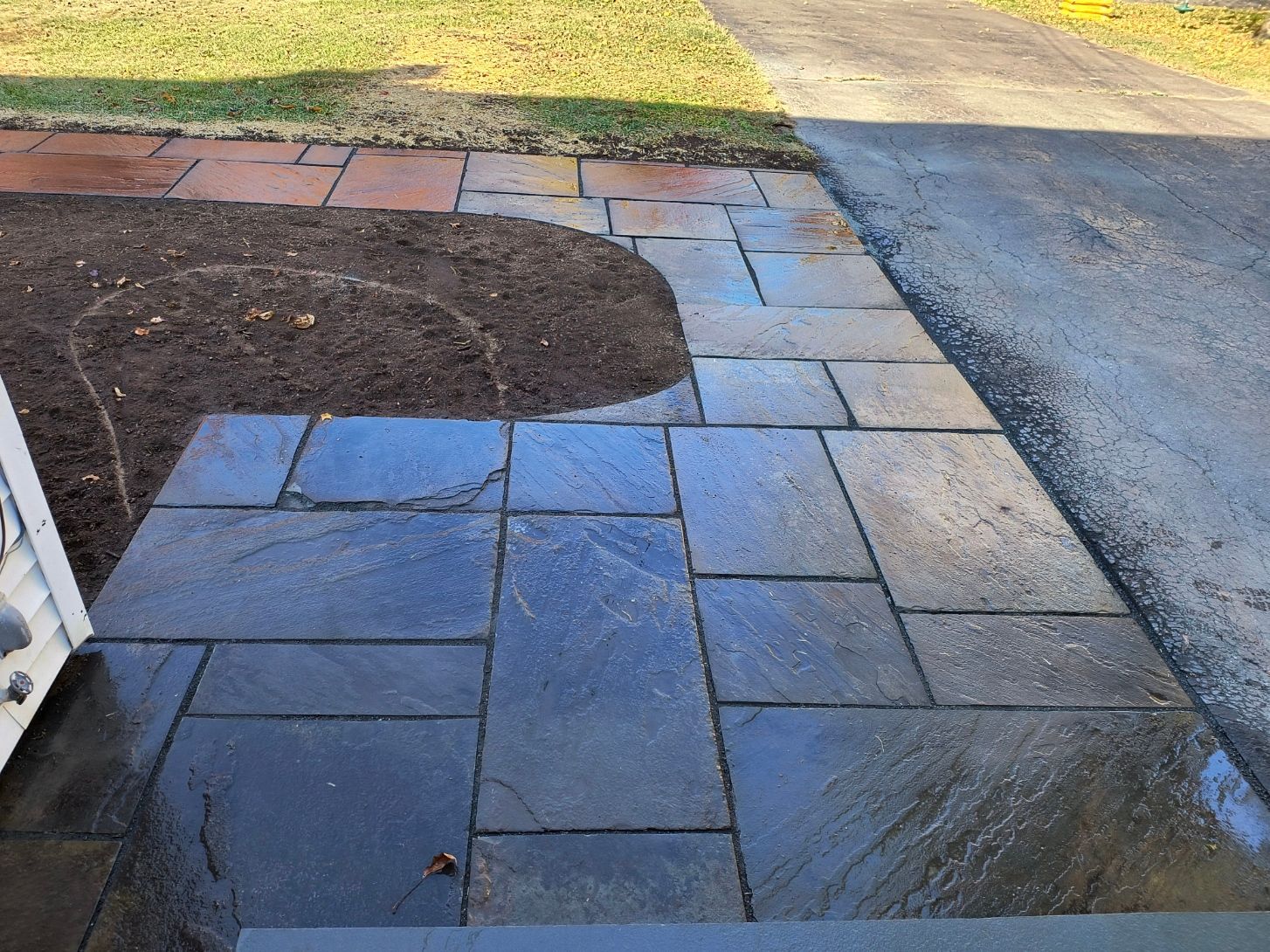 Stone walkway leading to a house entrance. Newly planted soil bed with adjacent driveway.