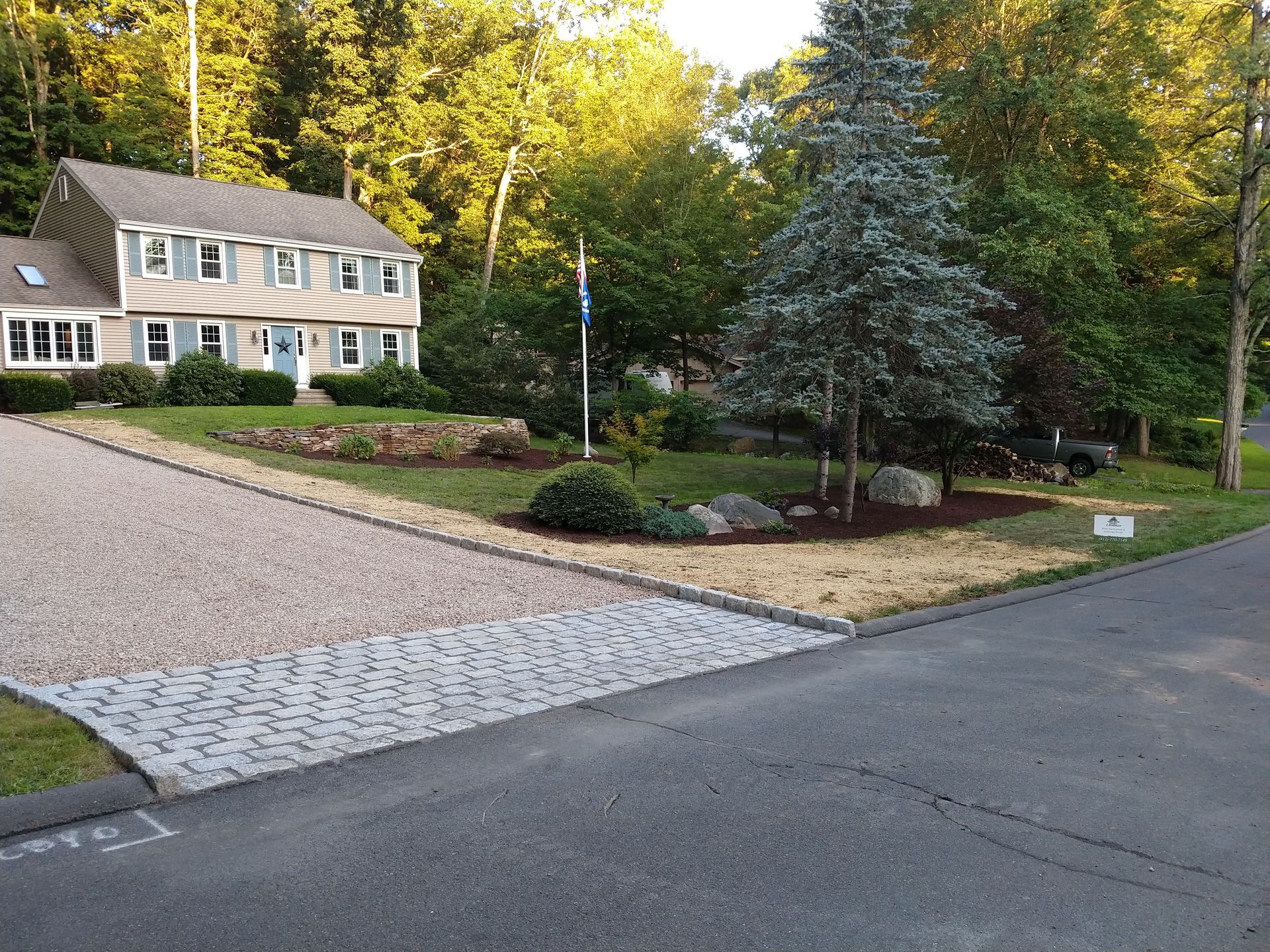 House with light-colored siding, gravel driveway, and paved border, trees in the background.