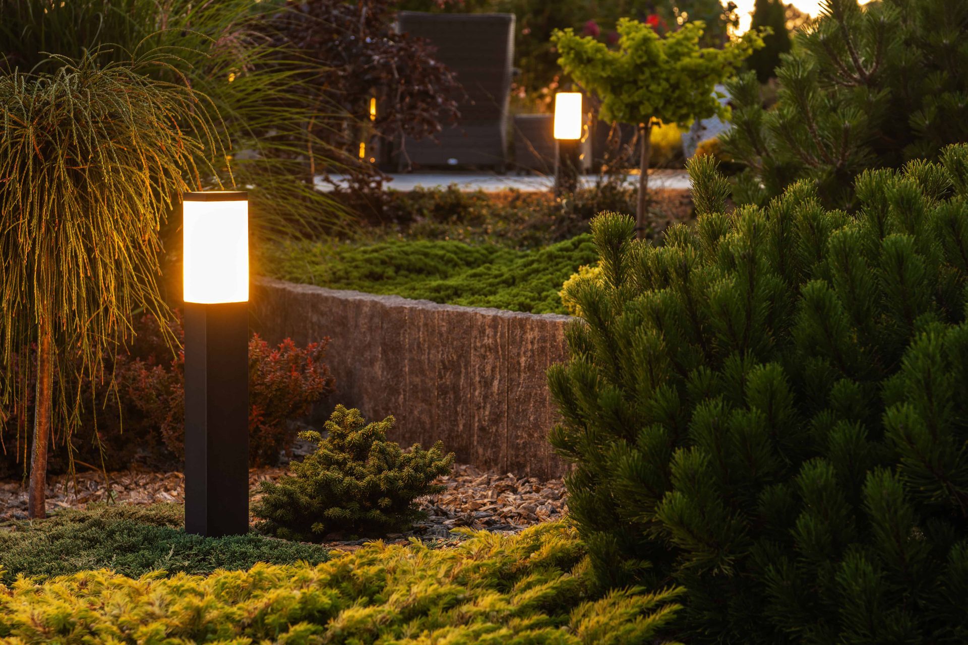 Lit garden with bushes, small trees, and rectangular pathway lights at dusk.