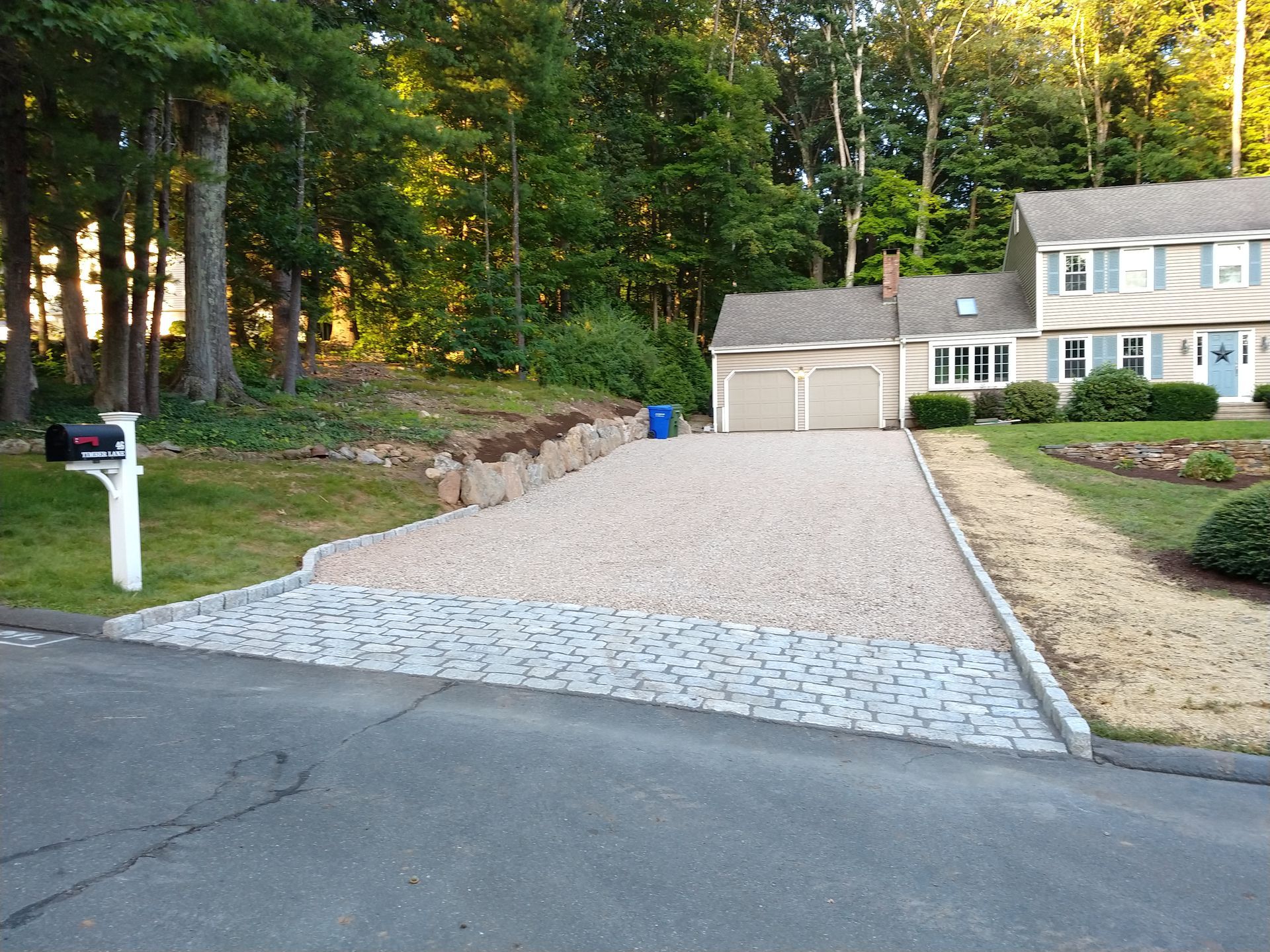 Driveway of a two-story house. Gravel driveway leads to a garage. Blue recycling bin and mailbox on the left.