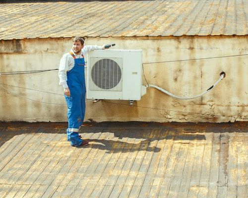 A man in blue overalls is standing next to a wall mounted air conditioner.