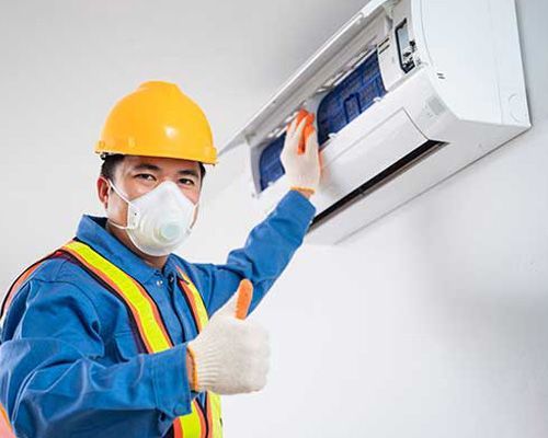 A man wearing a mask and hard hat is cleaning an air conditioner and giving a thumbs up.