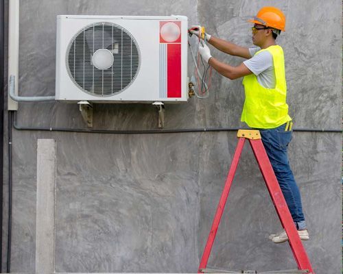 Arctic Fox technician is standing on a ladder fixing an air conditioner on the side of a building.
