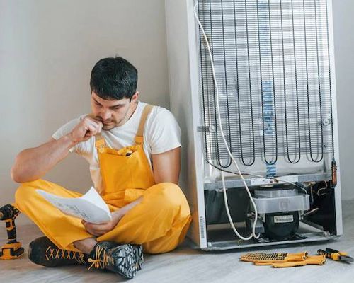 Arctic Fox technician in yellow overalls is sitting on the floor next to a refrigerator.