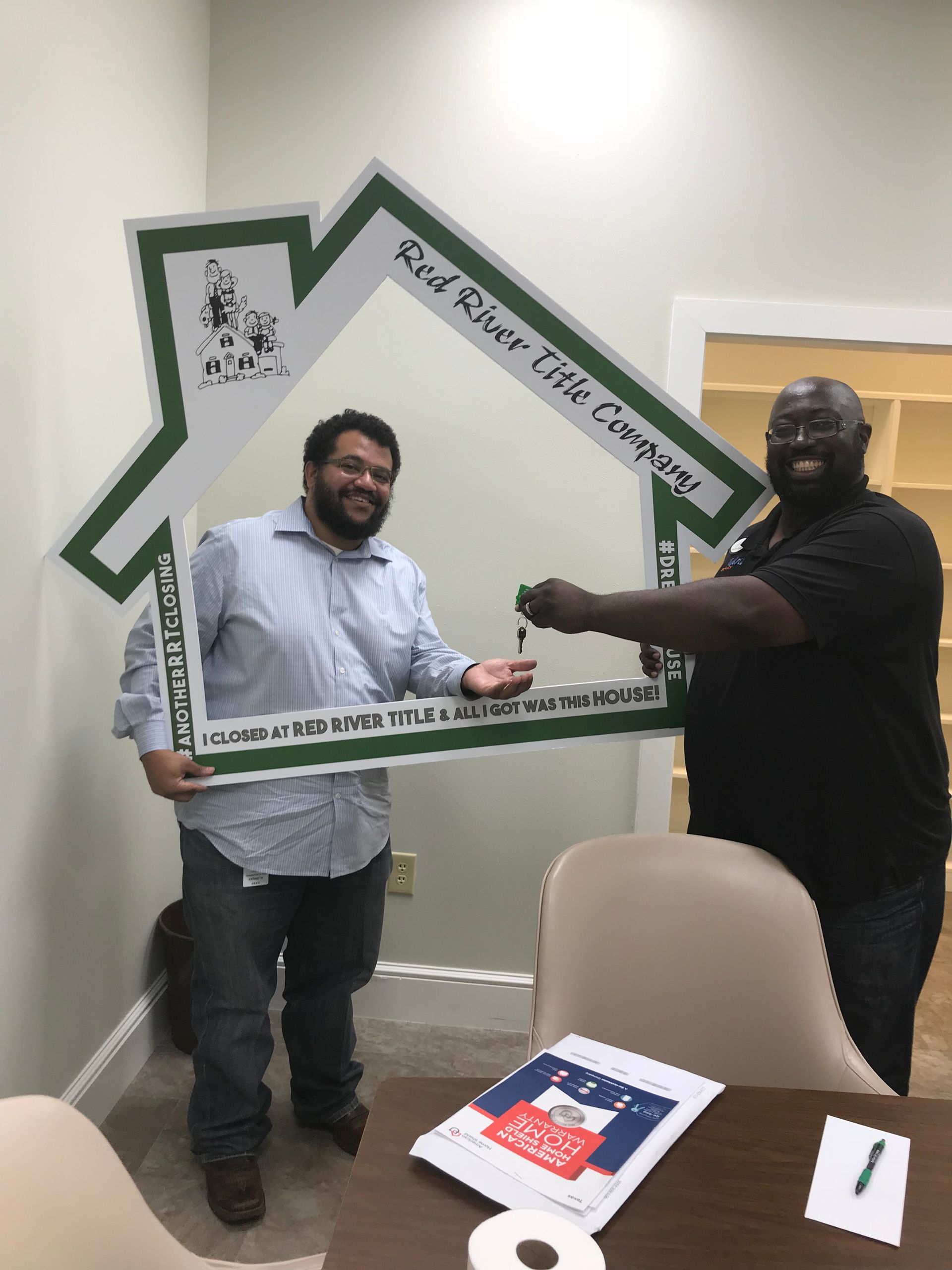 Two men hold a house-shaped frame and a key. One smiles, the other appears to hand off the key.