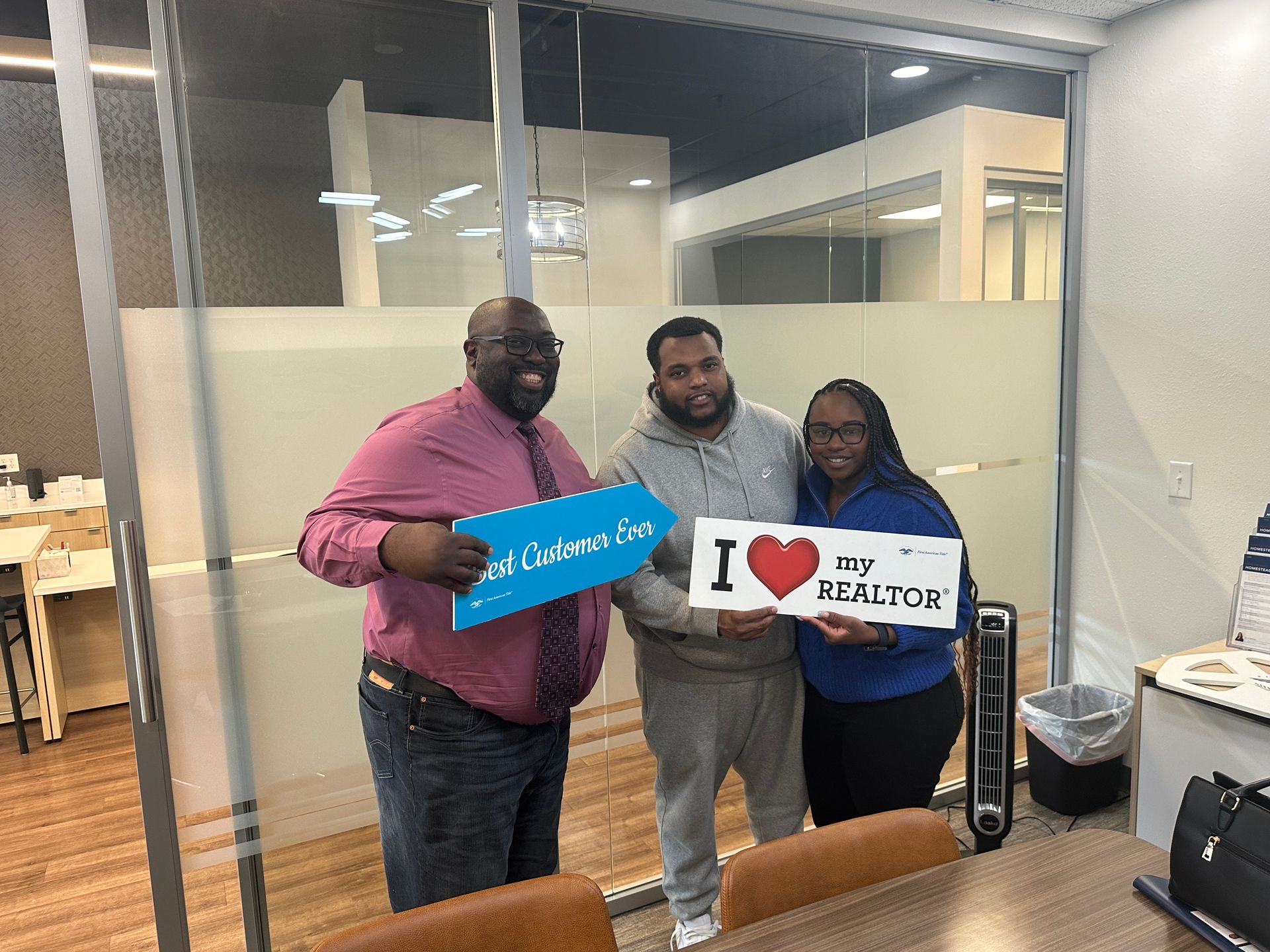 Three people pose with signs in an office. One holds 