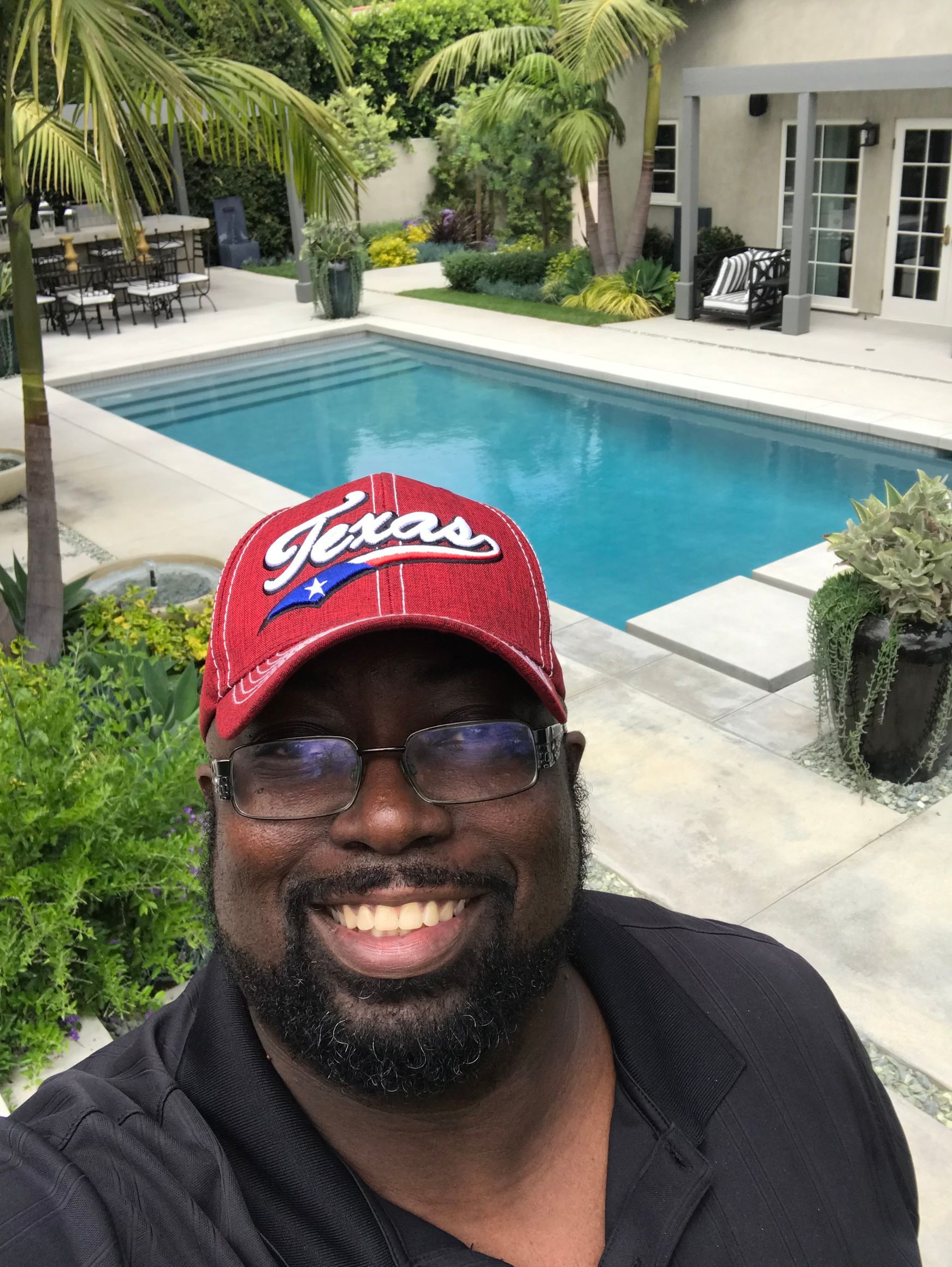 Man in Texas hat smiles in front of a pool, patio, and lush landscaping.
