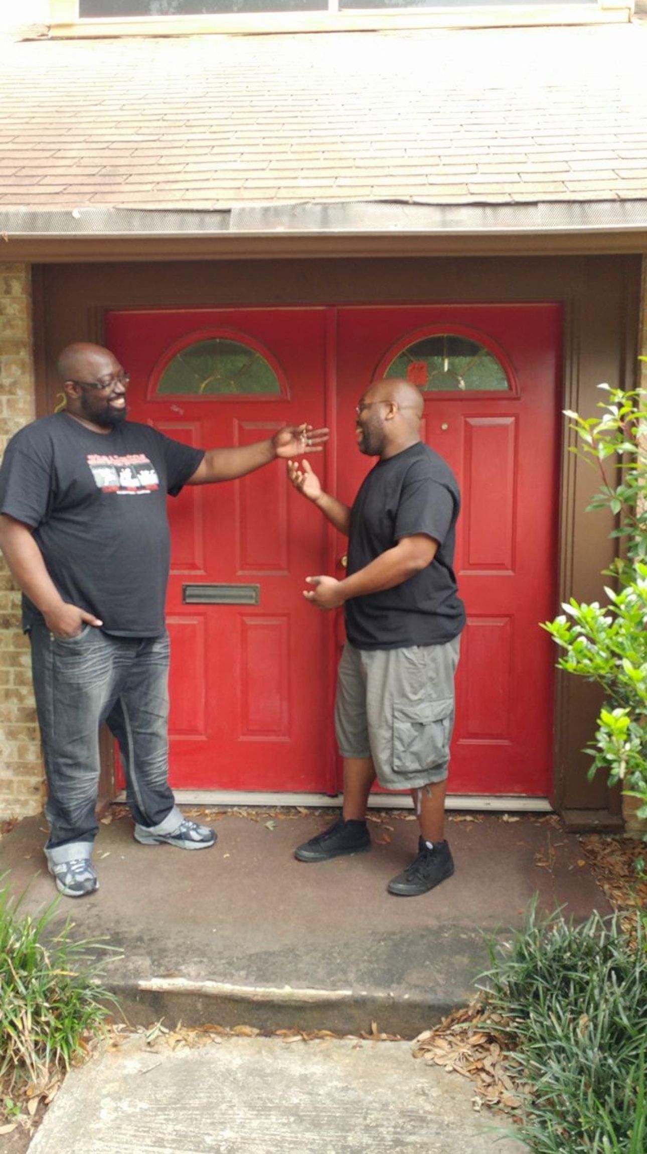 Two men standing by a red door; one points, the other gestures in conversation. Outdoors.