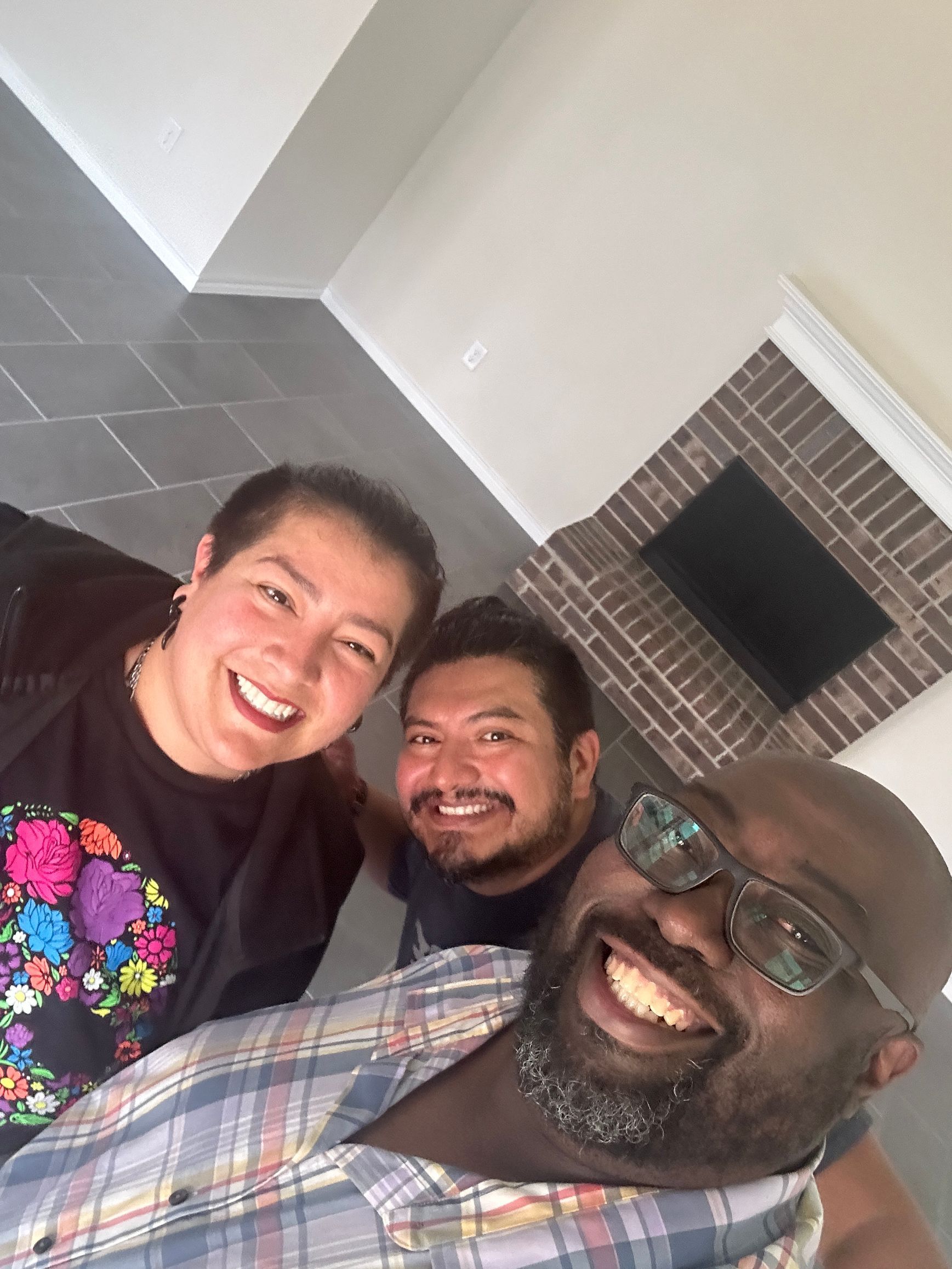Three smiling people taking a selfie; two men and a person with short hair; in a room with a brick fireplace.