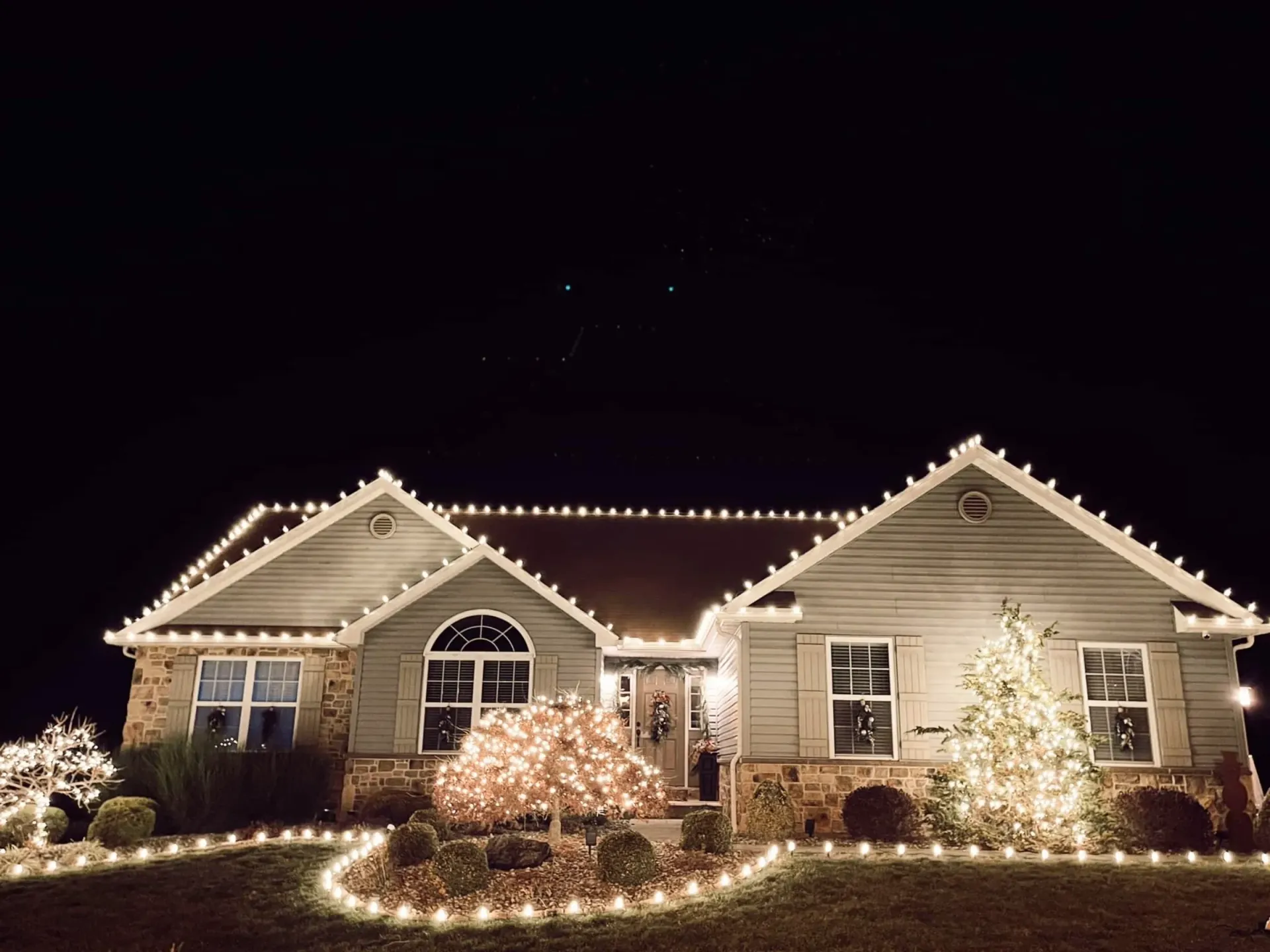 House decorated with warm white Christmas lights at night, centered in a dark sky.