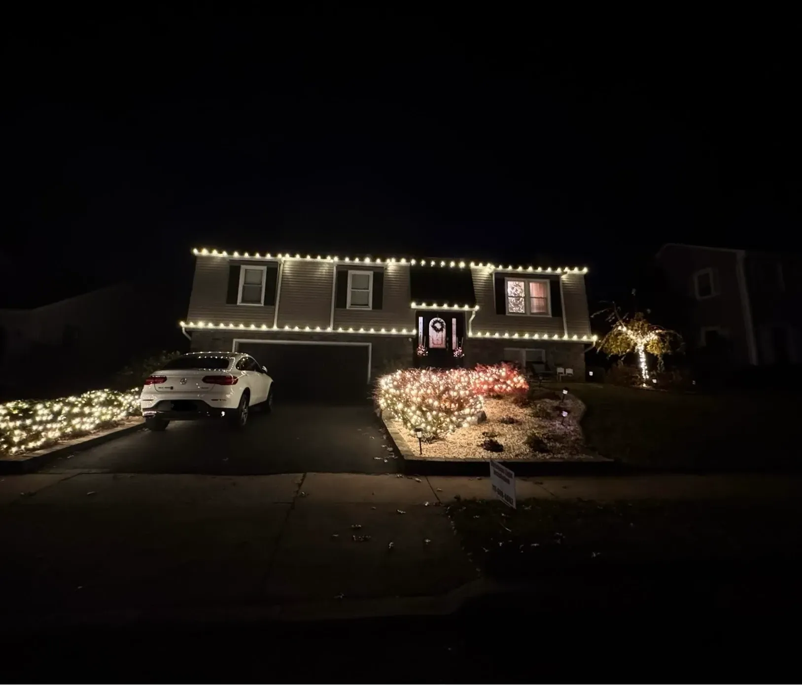 House decorated with white lights and illuminated holiday decorations at night, with a car in the driveway.