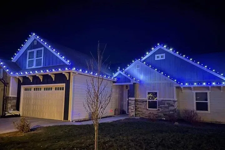 Suburban houses at night outlined with blue Christmas lights, one with a lit garage.