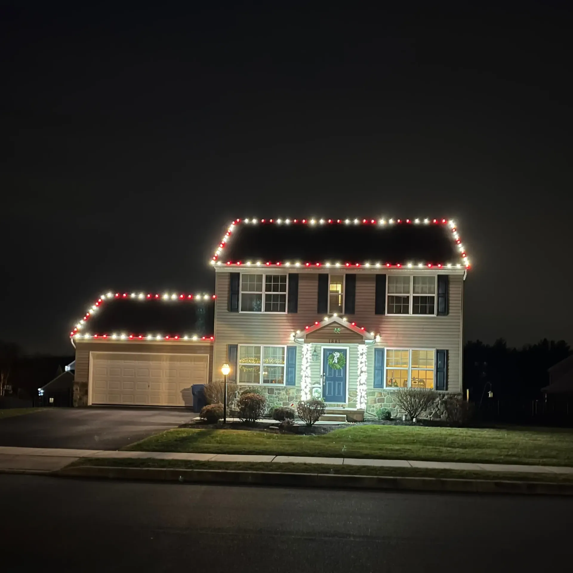 Two-story house at night, outlined in red and white Christmas lights with a lit blue front door.
