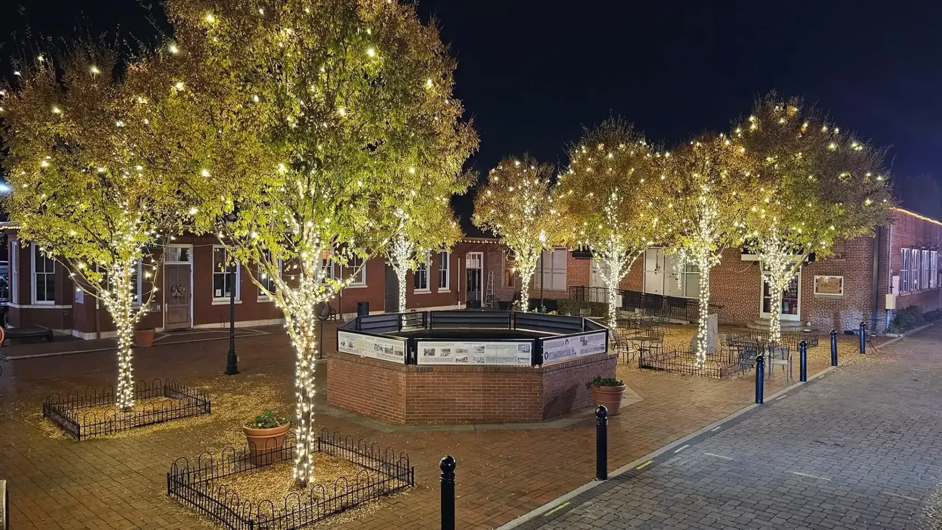 Nighttime plaza with lit trees, benches, and a cobblestone walkway beside buildings