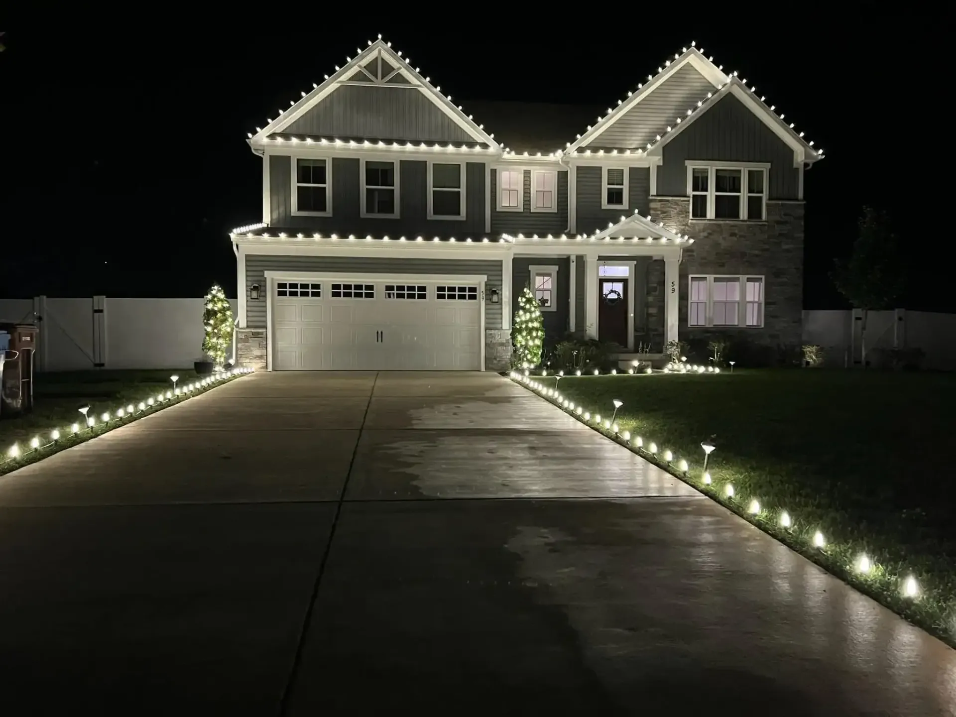 Two-story house at night with white holiday lights outlining the roof and a lit driveway