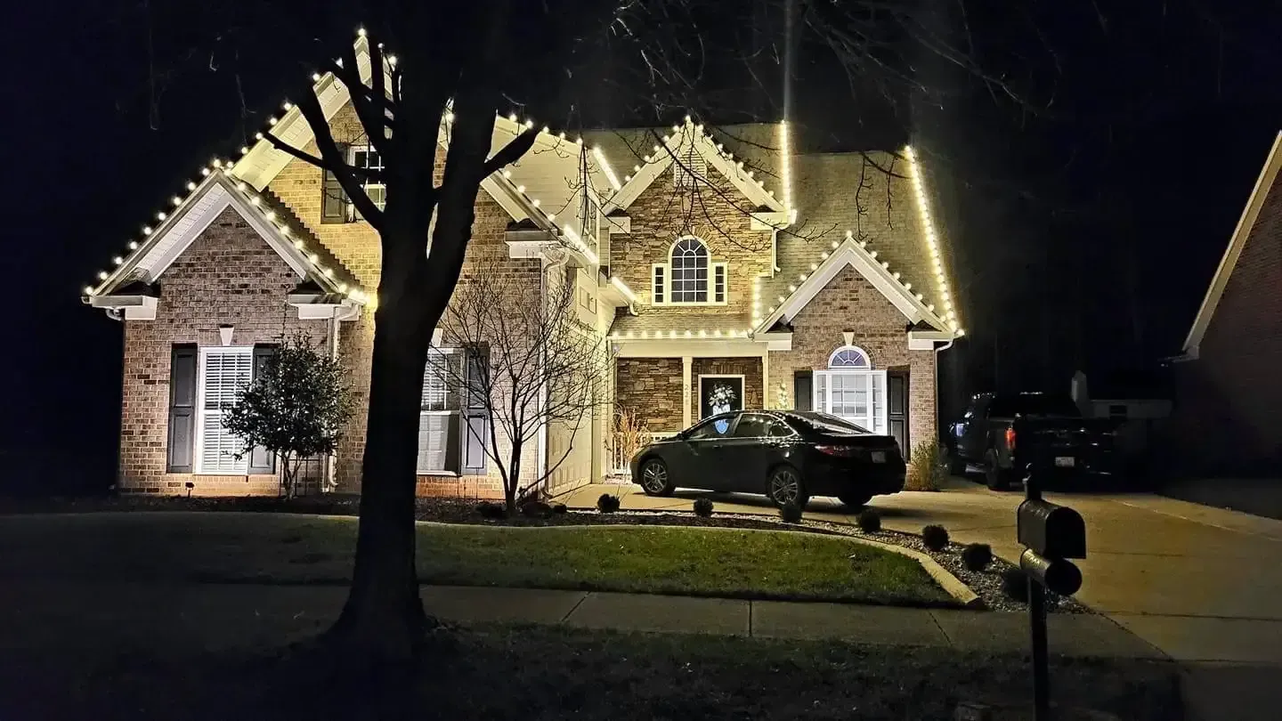 Nighttime view of a lit suburban house with a parked car, trees, and a lawn.
