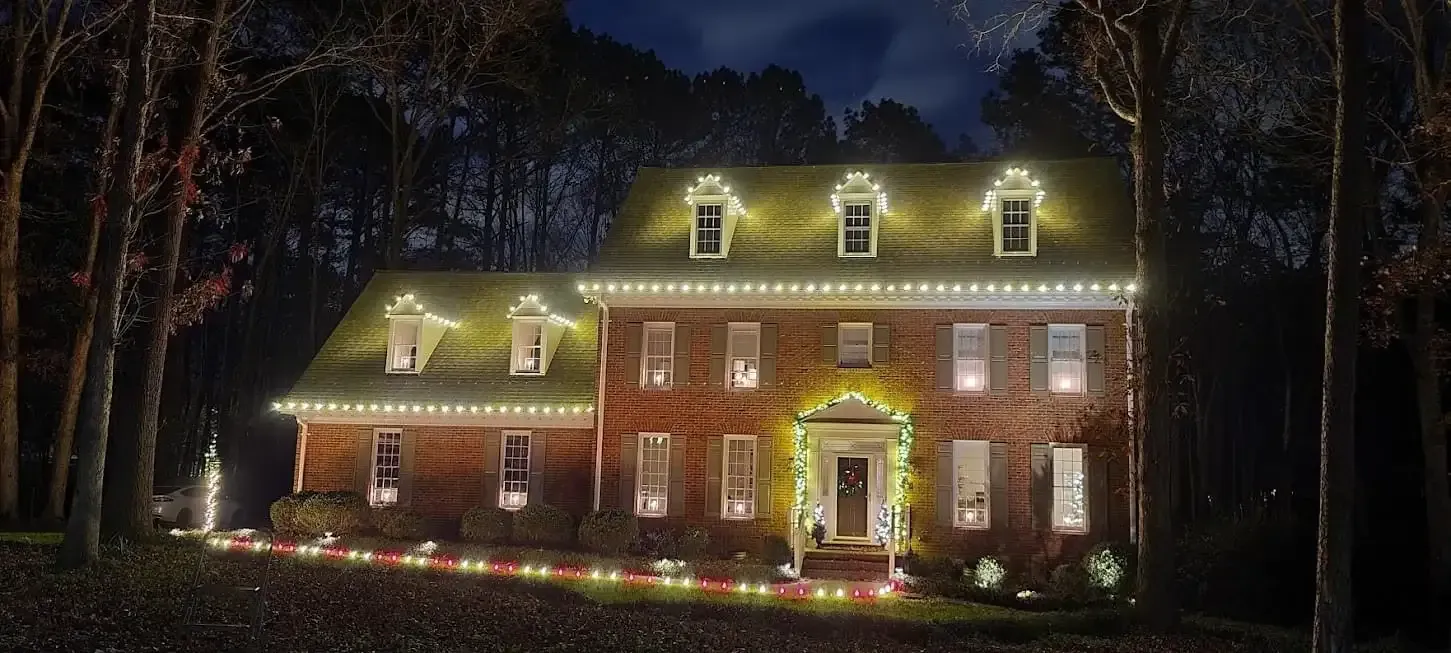 Brick house at night outlined with warm white holiday lights in a wooded setting