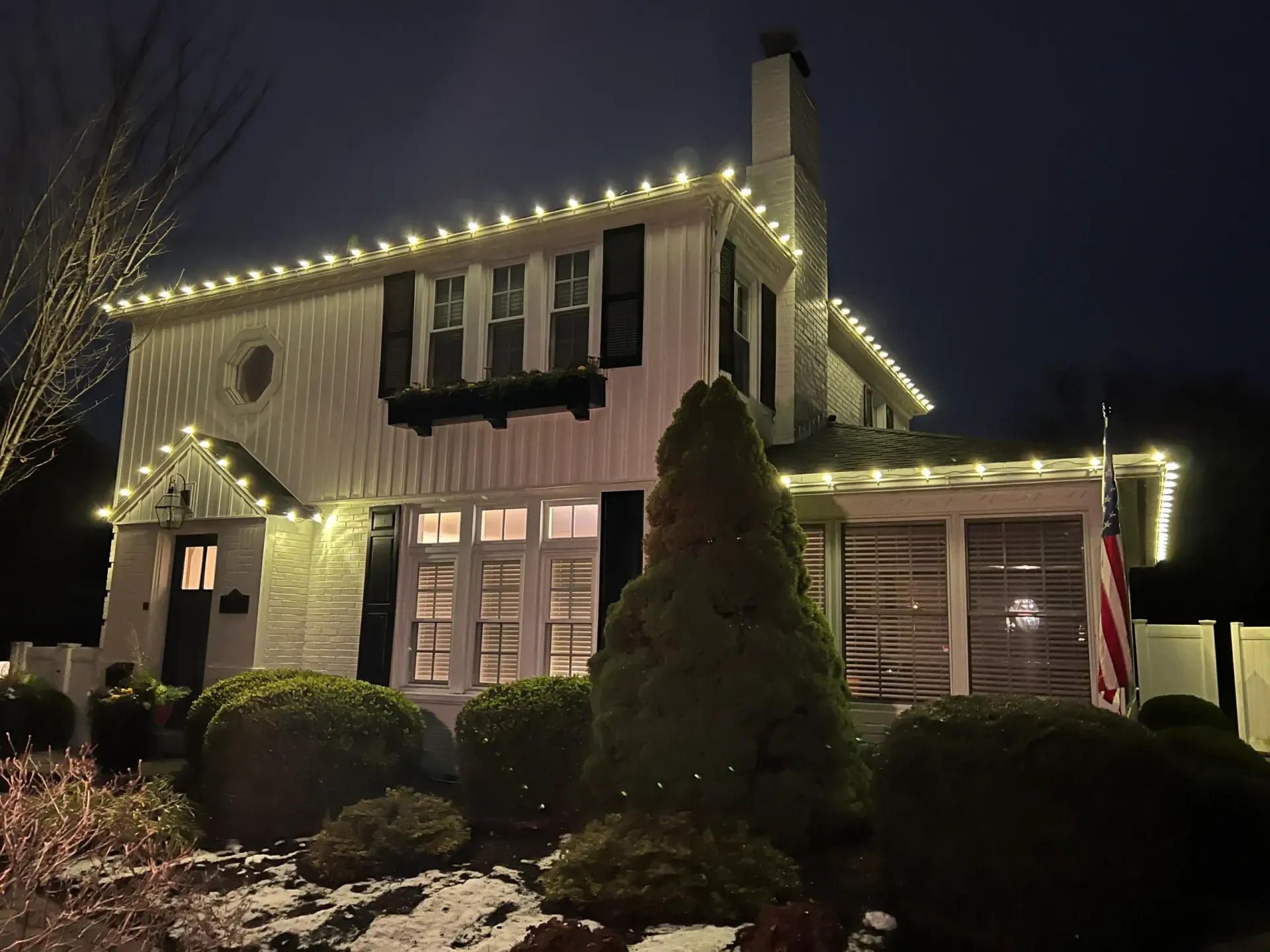 House decorated with warm white holiday lights at night, with shrubs and a lit chimney.