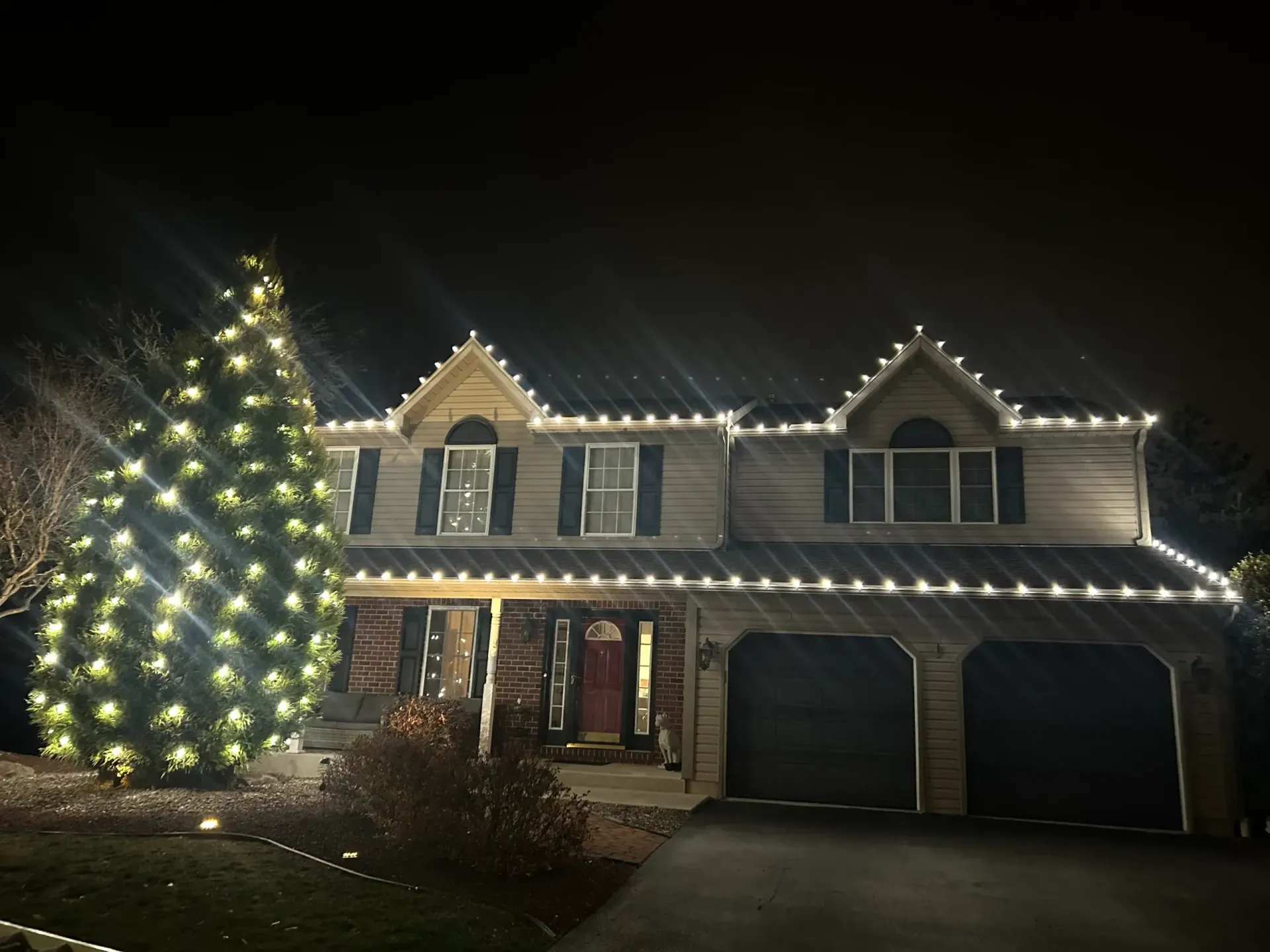 Two-story house at night with a lit Christmas tree and roofline lights, garage doors visible