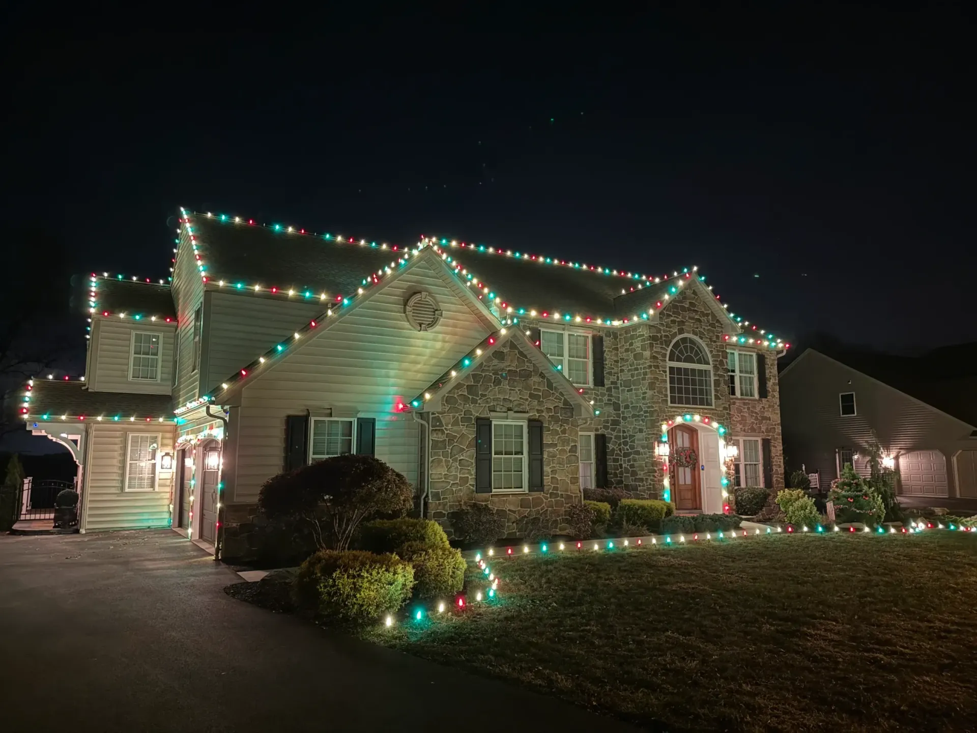 House decorated with colorful Christmas lights at night, with lit shrubs and a driveway.