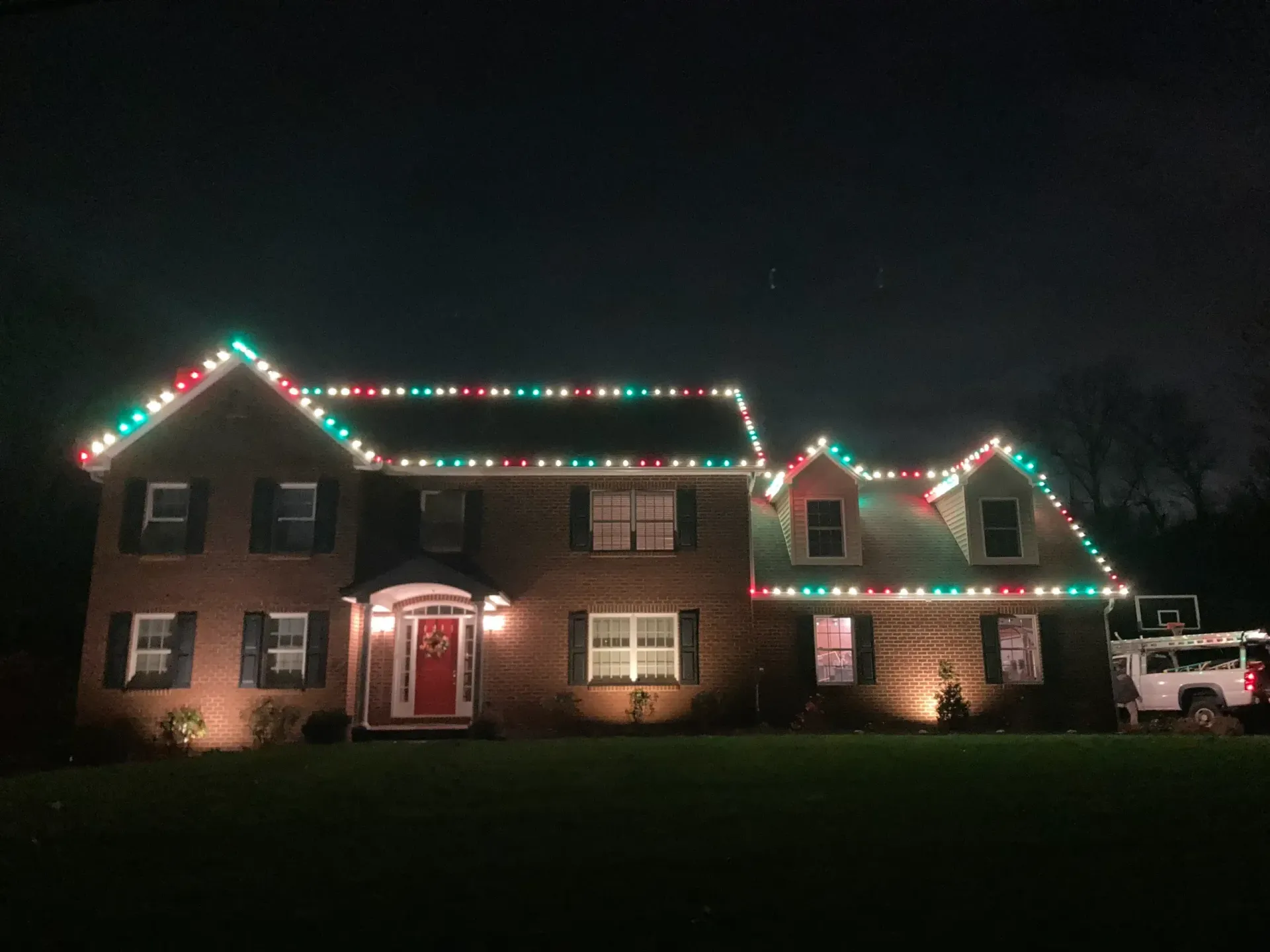 Brick house at night outlined with red, green, and white Christmas lights, with a lit front door and parked truck.