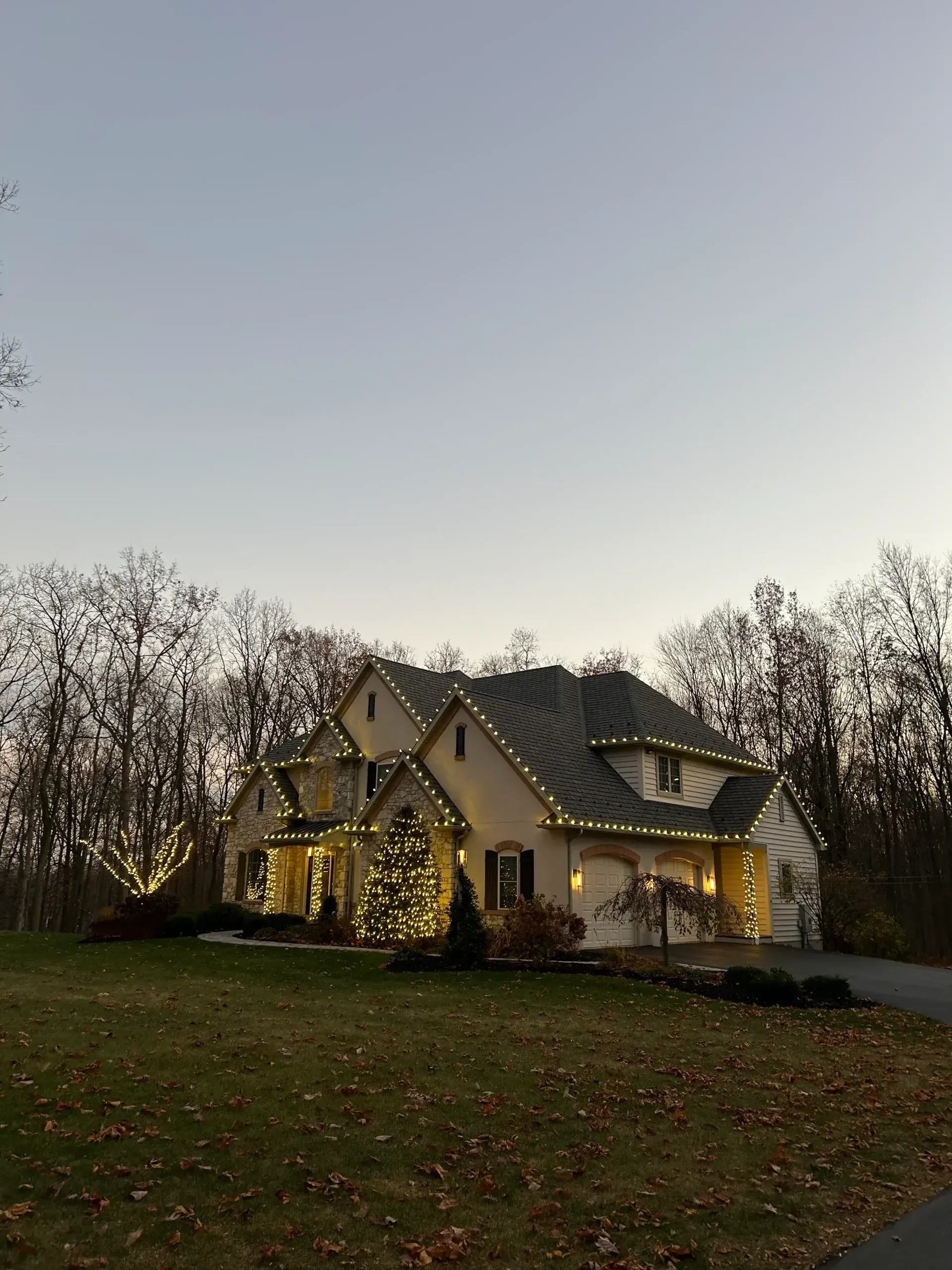 Lit house with warm porch lights in a grassy yard at dusk, framed by trees.