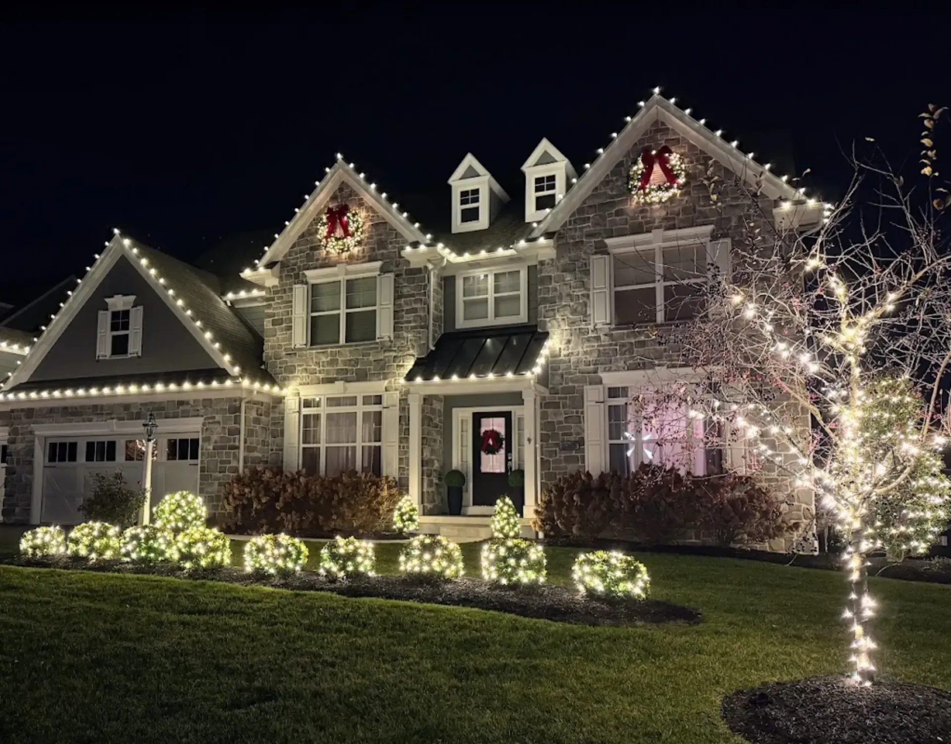 Two-story house decorated with white holiday lights, with lit shrubs and a glowing tree at night