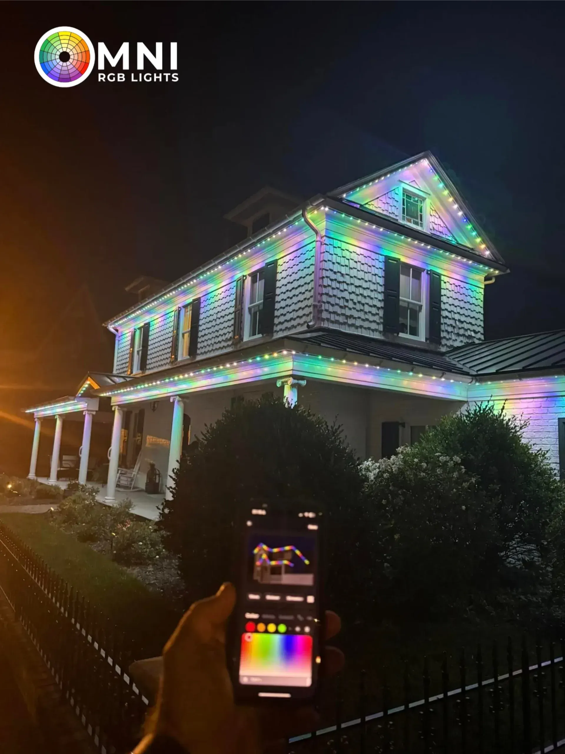 House covered in colorful holiday lights at night, with a hand holding a phone controlling the display.