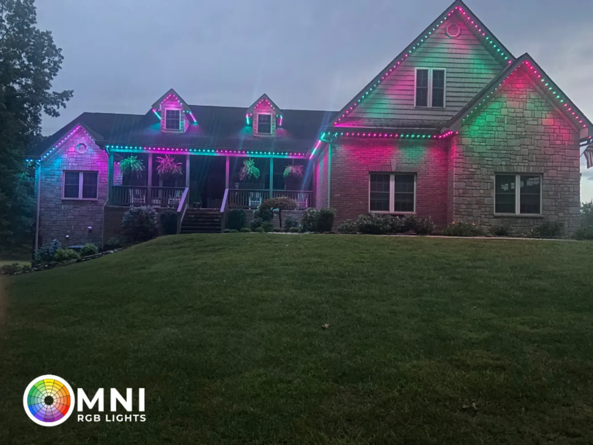 Stone house lit with pink and green holiday lights above a dark lawn at dusk