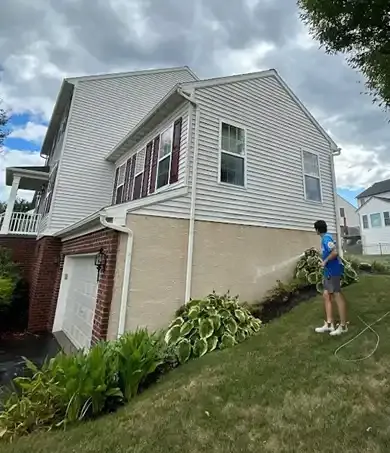 Person standing on a sloped lawn beside a two-story house with a garage, holding a hose.