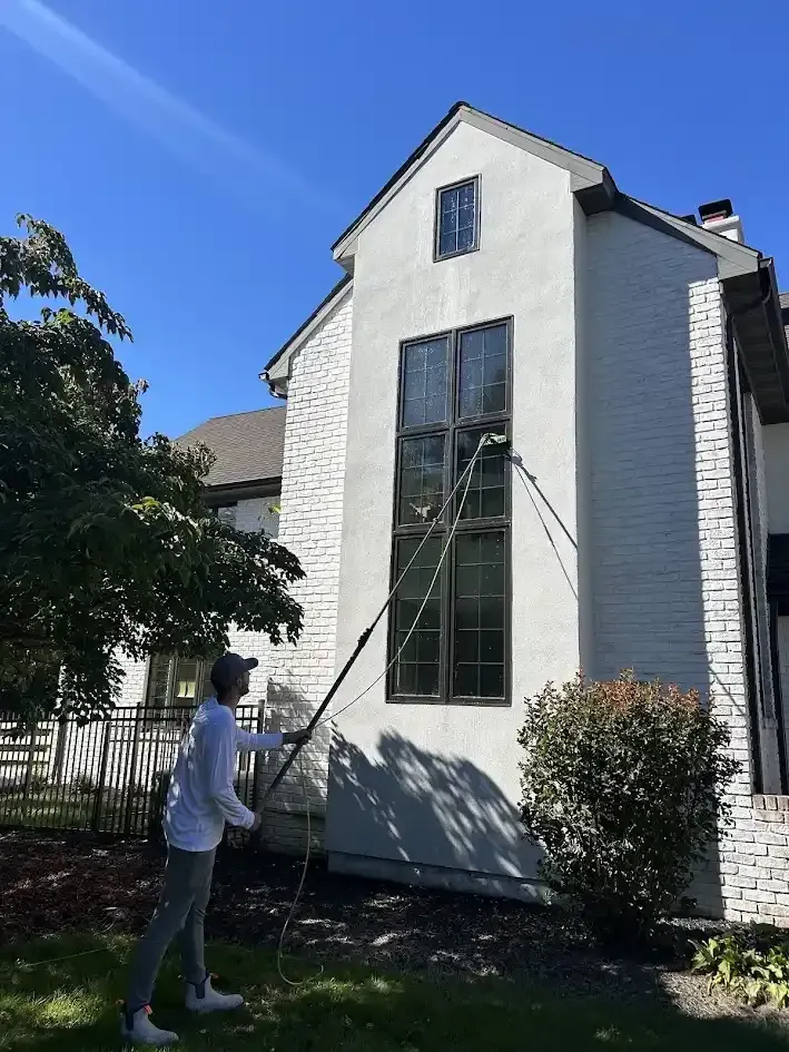 Person using a long pole to clean a second-story window on a white house.