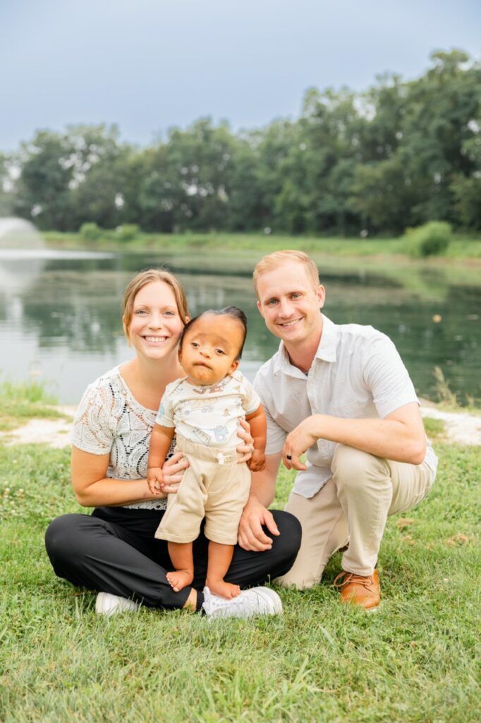 Smiling family of three posing on grass by a lake with trees in the background
