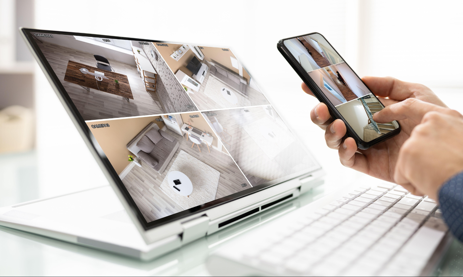 Hand holding smartphone beside a laptop showing a home security camera feed