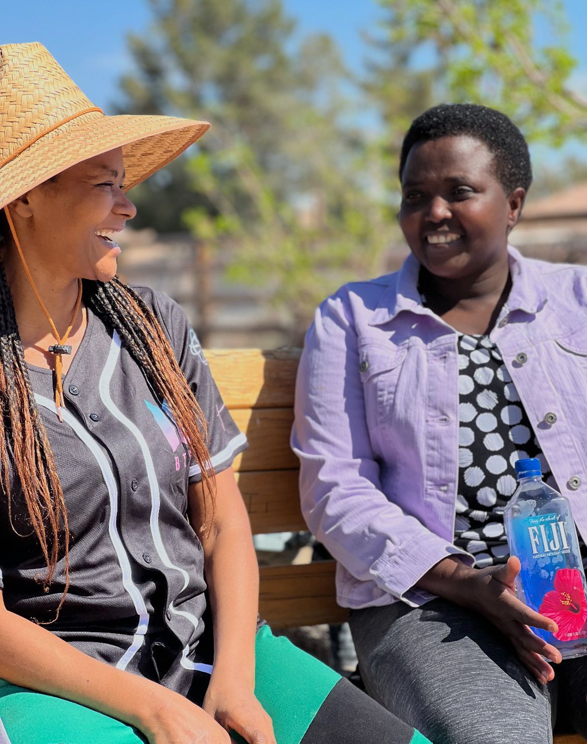 Two women outdoors, laughing. One in a straw hat, the other in a purple jacket holding Fiji water.