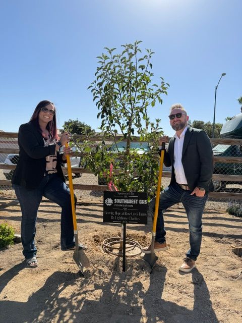 Two people standing beside a newly planted tree with a commemorative sign on a sunny day.