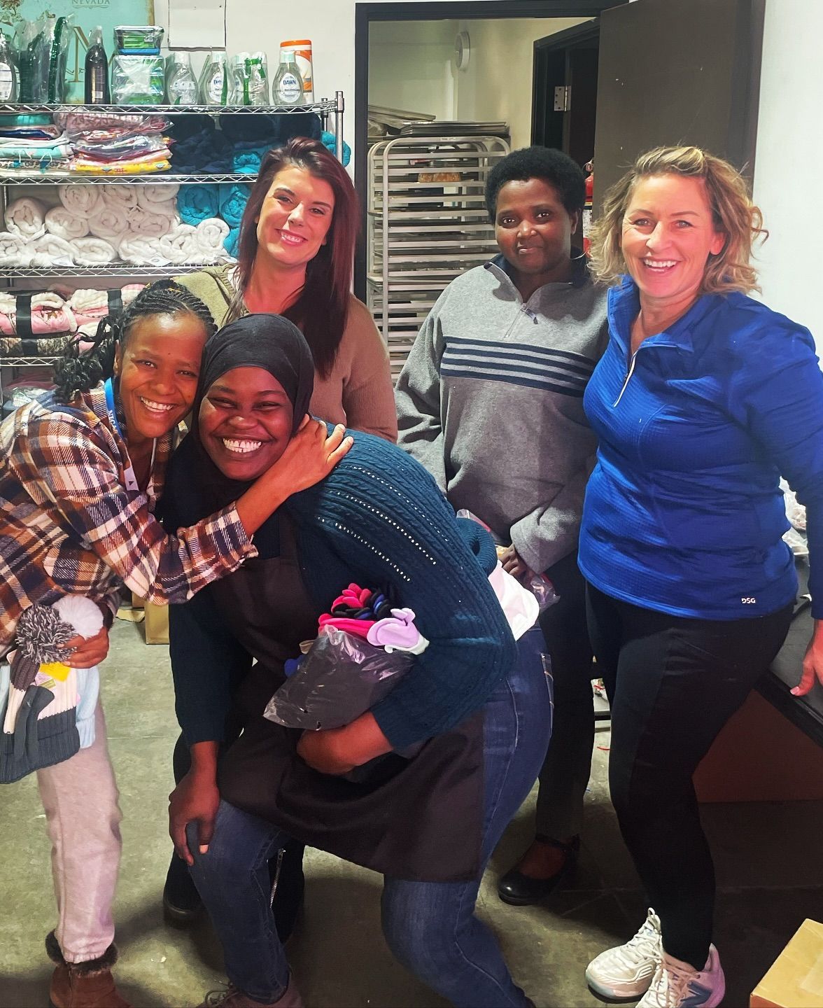 Five people, diverse in appearance, smiling and embracing in a brightly lit commercial kitchen setting.