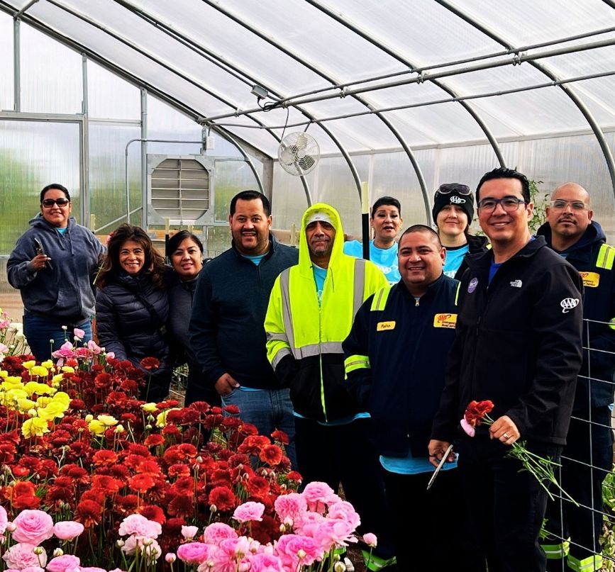 Group of people smiling in a greenhouse with colorful flowers; some holding flowers.
