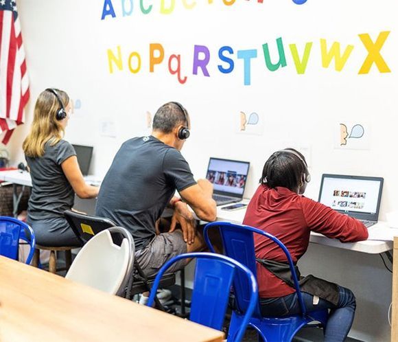 Three people wearing headsets sit at laptops in a classroom with alphabet letters on the wall.