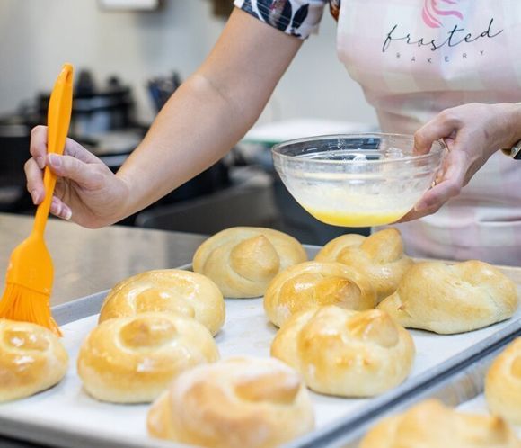 A baker brushing glaze on baked buns on a tray. Frosted Bakery apron visible.
